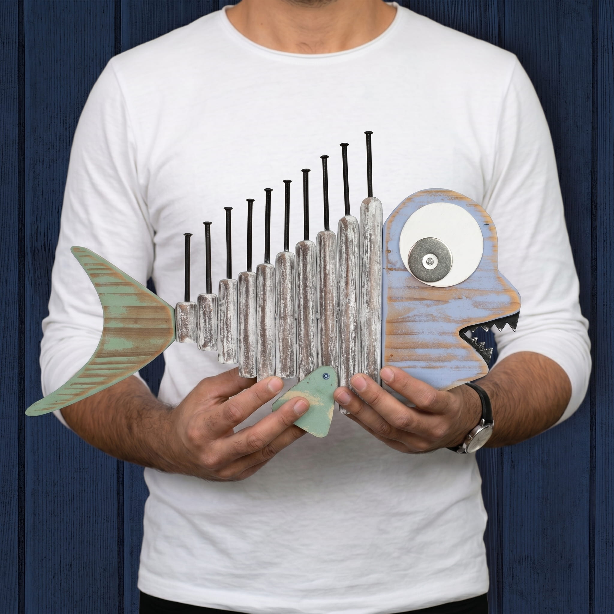 A person wearing a white long-sleeved shirt holding a piece of handcrafted wooden fish wall art, featuring textured driftwood-style wood and nail spines against a dark blue plank-style background